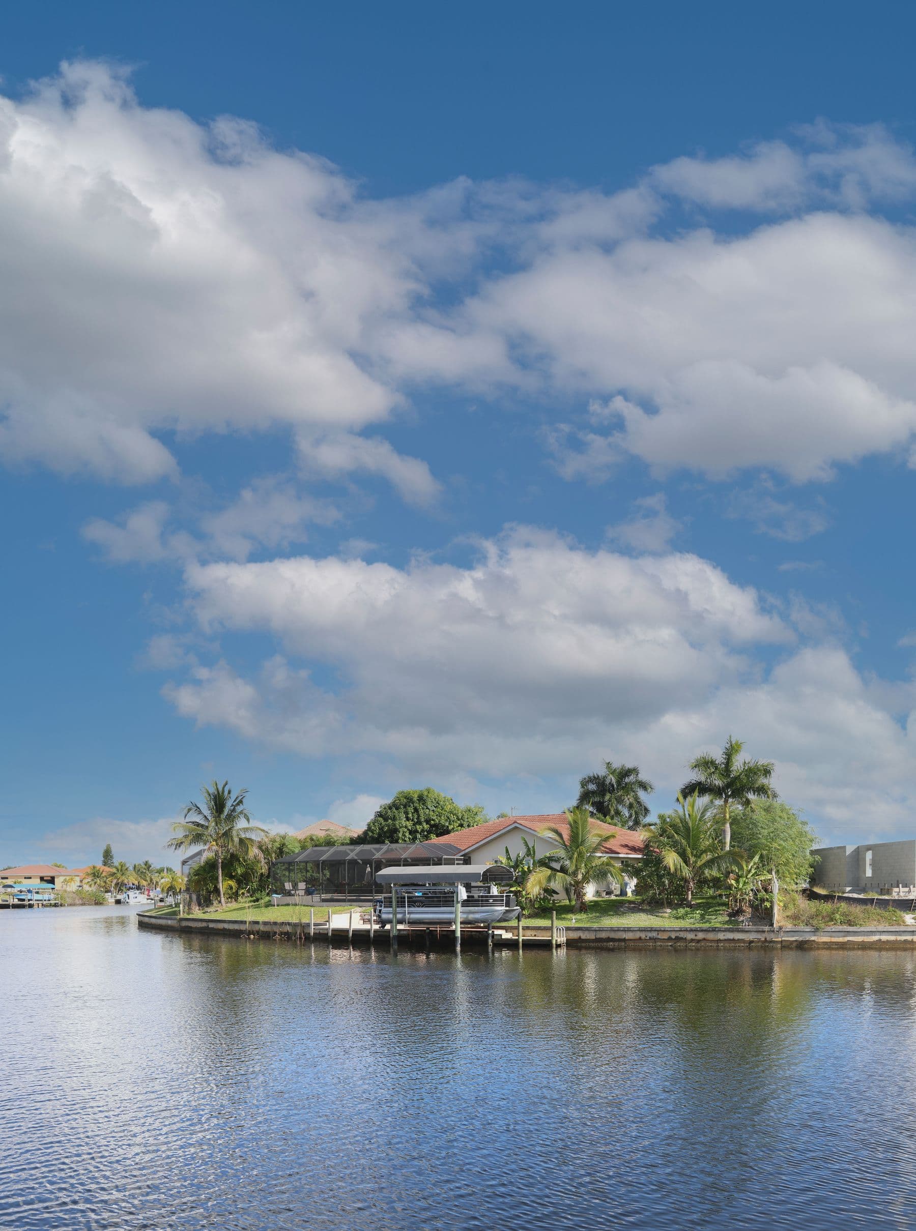 A single-story house with a red tile roof, a screened pool cage and a boat lift on a canal under a large cloudy sky.