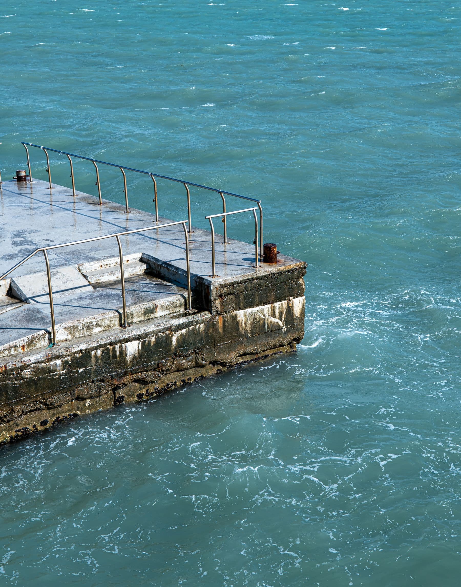 A weathered concrete pier with steel railings and a rusted bollard reaching into choppy blue-green water.