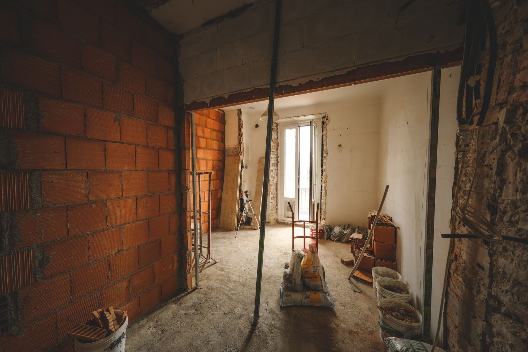 A room stripped to bare brick and concrete, with a hand truck, buckets and bags of material on the floor.