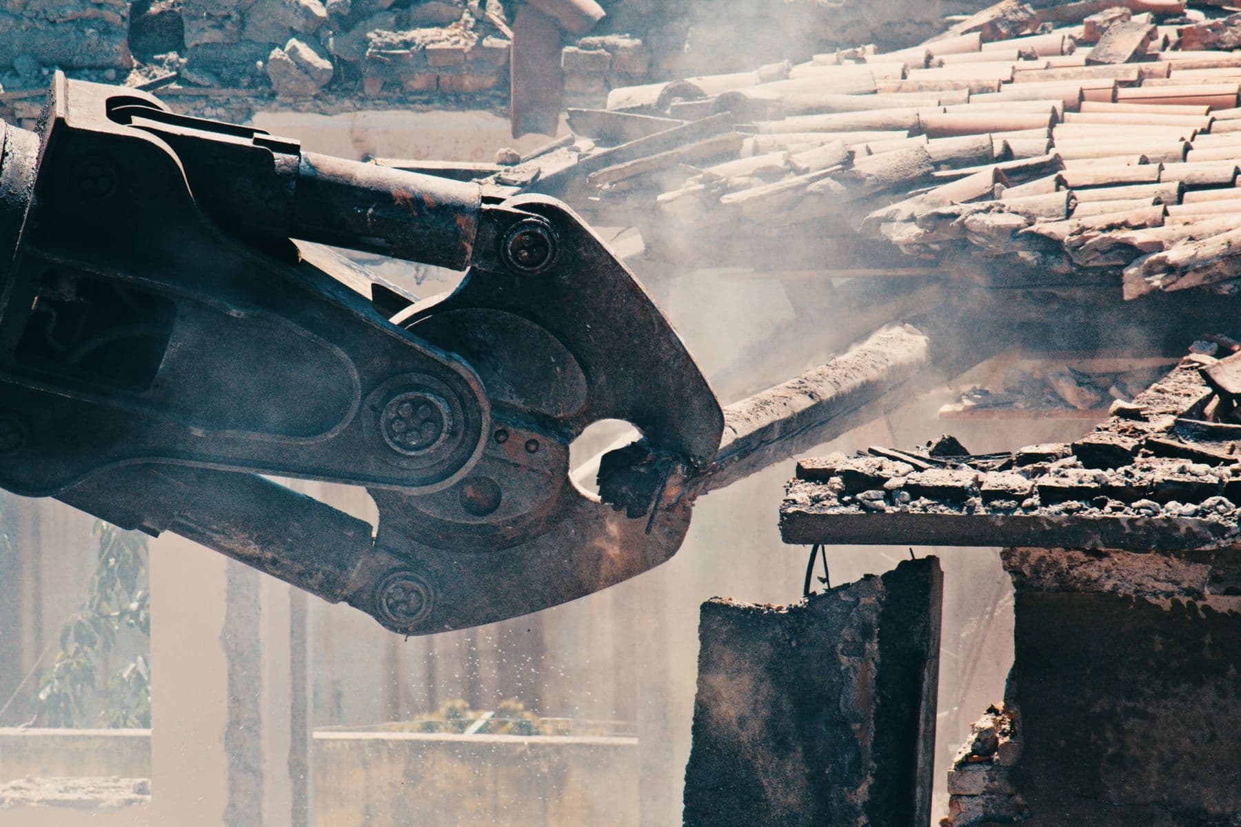 The steel jaws of a demolition shear gripping a beam beside a clay-tile roof, with dust in the air.
