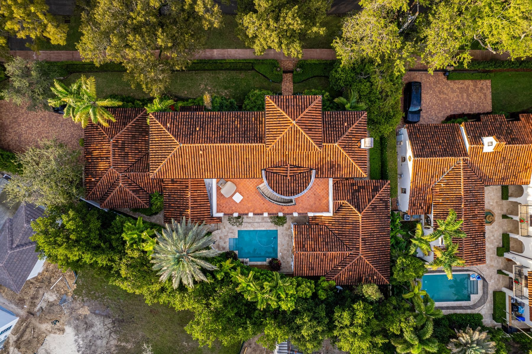 An overhead view of terracotta tile roofs with two small backyard swimming pools among palms and trees.